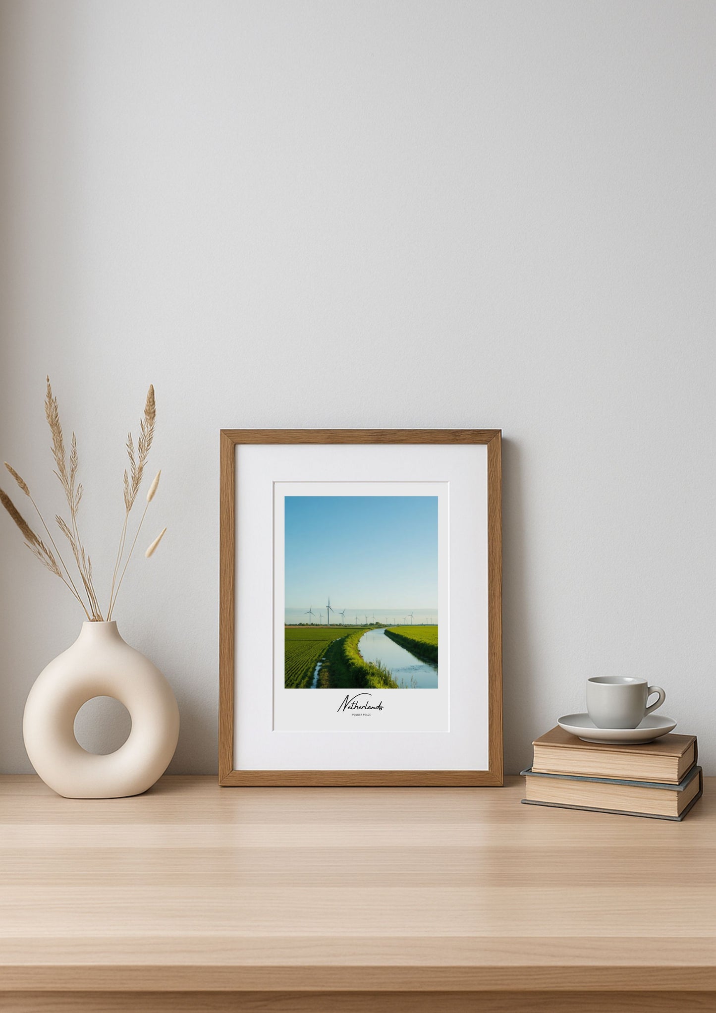 Framed artwork on a wooden shelf with a vase and books against a white wall