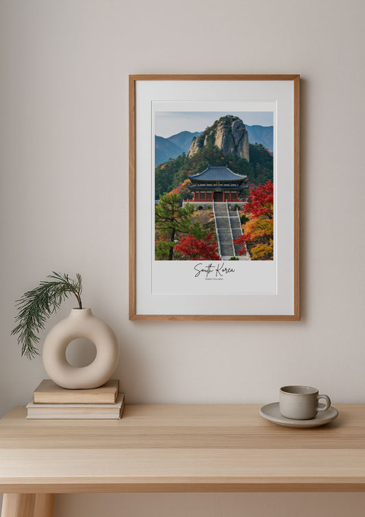 Framed print of a scenic landscape with a building and mountains on a wall above a wooden table.