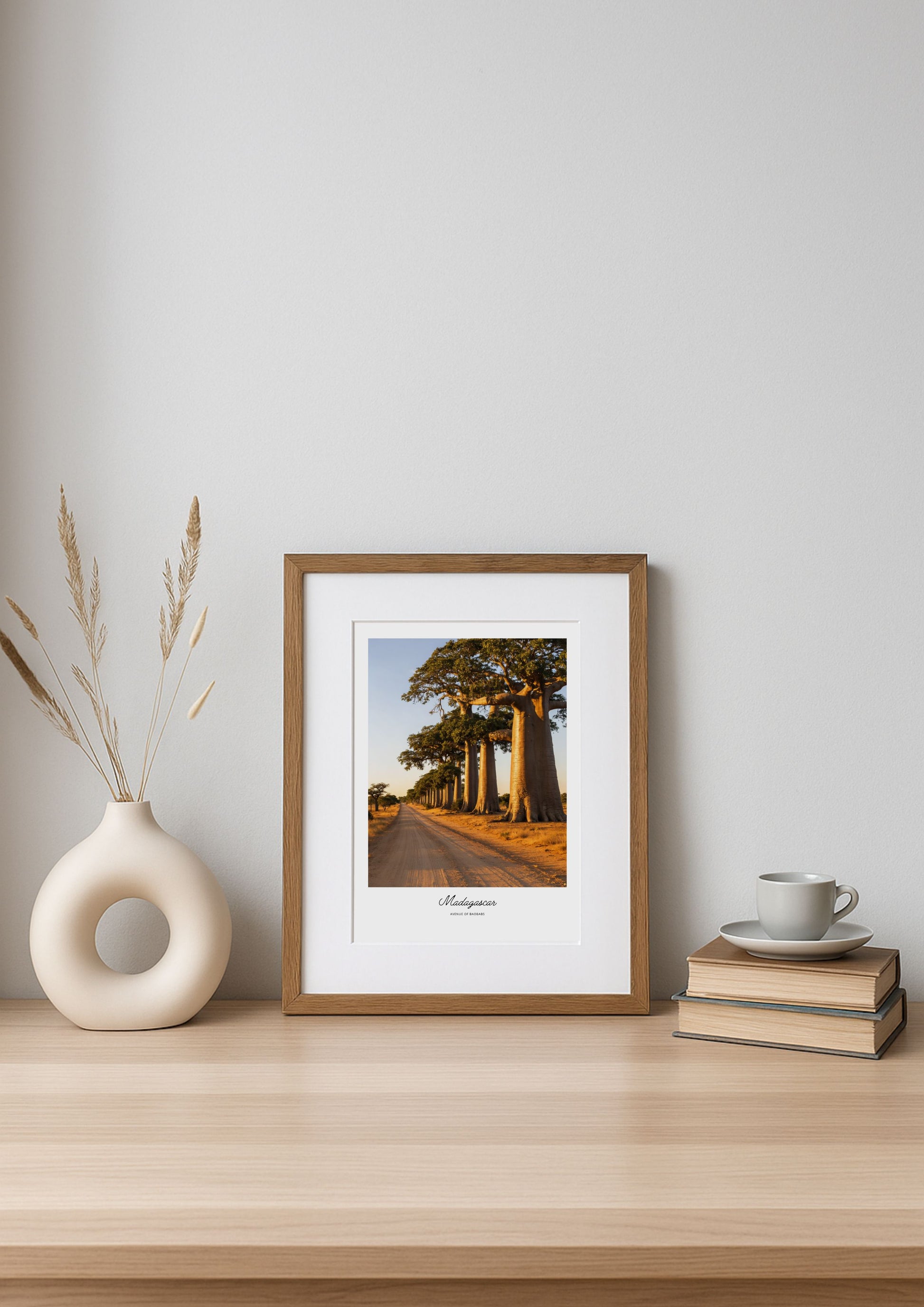 Framed artwork on a wooden shelf with a vase and books against a white wall