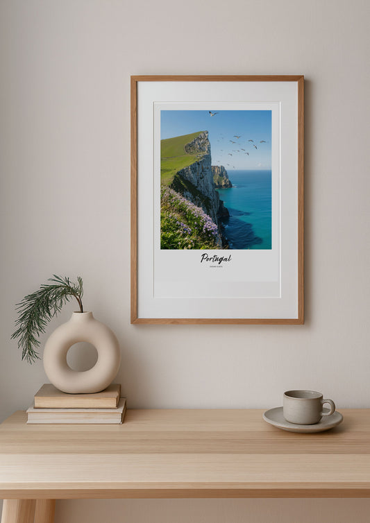 Framed artwork of a coastal scene with cliffs and sea, placed on a wall above a wooden table with decor items.