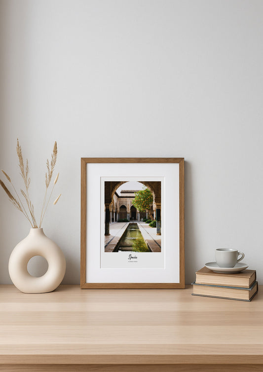 Framed artwork on a wooden shelf with a vase and books against a white wall