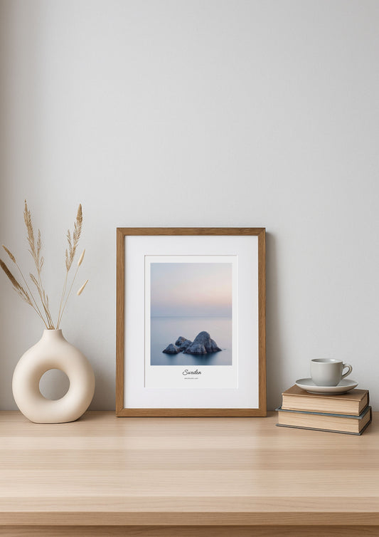 Framed artwork of a coastal scene on a wooden shelf with a vase and books.