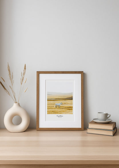 Framed artwork of a landscape with a deer on a wooden shelf against a white wall.